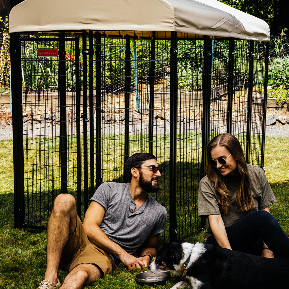 Smiling couple seated on grass beside an outdoor pet kennel, enjoying the day with their content dog.